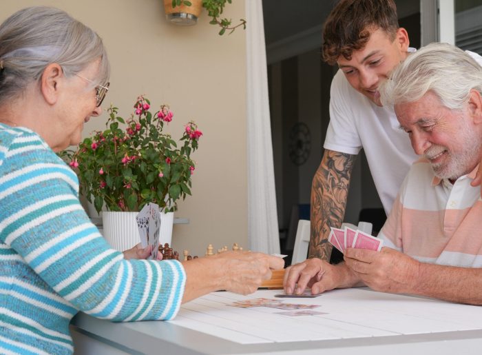 Happy multi generation family enjoying free time together sitting outdoors on terrace smiling playing cards. Grandparents and young grandson stay together in leisure activity